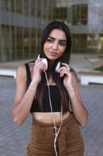 A young woman with long dark hair holds white headphones around her neck while standing outside.