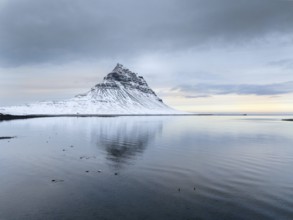 A peaceful winter scene of Kirkjufell Mountain in Iceland, covered in snow. The mountain reflects
