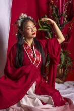 A woman in a red robe sits on a bed with a potted plant behind her. She is wearing a flower