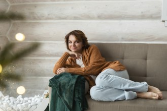 A woman in a cozy brown cardigan and blue pants lounges on a gray sofa, looking at the camera