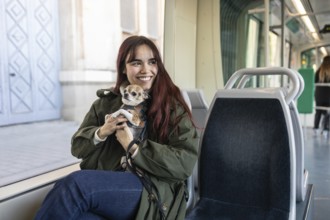 A cheerful woman sits on a tram seat holding her small chihuahua, warmly wrapped in a coat. The