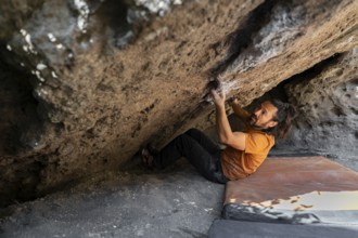 A climber in an orange shirt tackles a challenging boulder face in the wilderness, gripping rocks