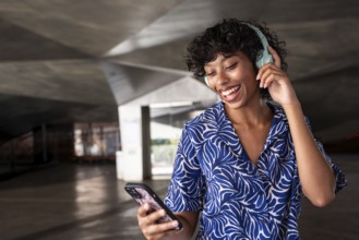 A joyful woman enjoys music through her headphones while holding a smartphone, standing in a modern