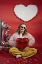 A young woman smiles joyously while holding a red heart-shaped balloon. She wears heart-shaped