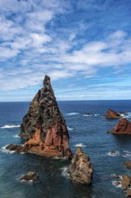 Steep rock formations rise out of the deep blue sea under a partly cloudy sky