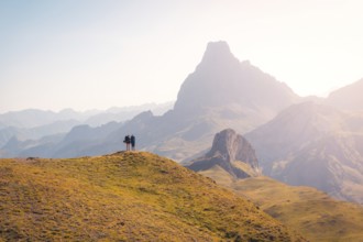 Two hikers stand on a grassy hill, gazing at the breathtaking mountain range in the Pyrenees. The