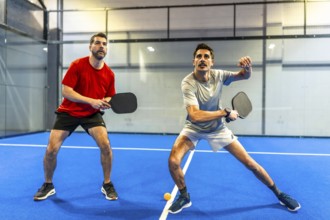 Two active men engaging in a pickleball match on a blue indoor court, concentrating on hitting the