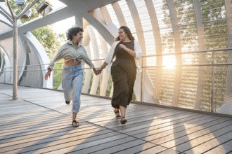 Lesbian couple joyfully holds hands while running together on a modern walkway, surrounded by
