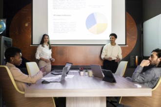 A diverse team engages in a modern office meeting. Two presenters stand by a screen displaying a