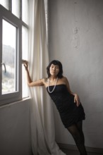 A stylish portrait of an Asian woman in a black dress, standing by a window with natural lighting.