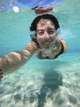 A woman swims underwater in Greece, capturing a selfie. The clear blue water reflects sunlight,