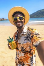 Young man taking a selfie on a tropical beach, wearing sunglasses and a straw hat, holding a