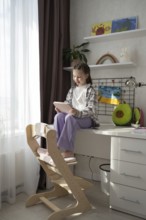A child sits on a desk, engaged with a tablet, surrounded by vibrant art, plush toys, and
