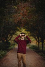 A man in a red sweater looks through binoculars amidst vibrant autumn foliage in a scenic park