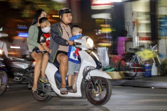 A family rides a scooter through a busy city street at night, people on scooters drive through the