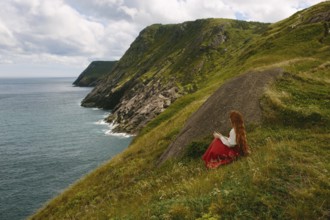 Side view of an unrecognizable redhead woman in a red dress, seated on a grassy slope reading a