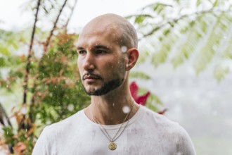 A man stands in the rain, deep in thought, surrounded by lush foliage in a forest. Drops fall