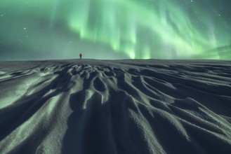 Unrecognizable person in red walks alone under the Northern lights on a snowy landscape at night,