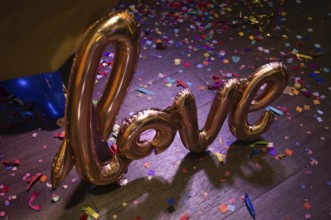 A golden foil balloon spelling love on a wooden floor covered in colorful confetti, creating a