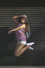 A young woman in sunglasses and a hat jumps joyfully against a dark urban background, embodying