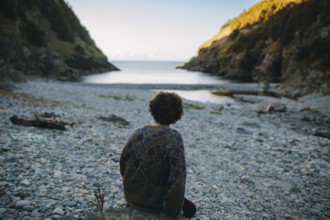 Back view of a man sitting contemplatively on a pebble beach, facing the tranquil sea as the sun