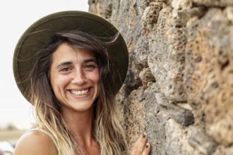 A woman wearing a hat smiles brightly while standing against a textured stone wall Her natural,