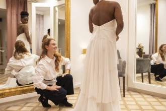 A bride to be wears a layered white gown, standing in a fitting room. Her designer, in a white