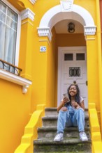 Vertical portrait of a smiling african woman using phone sitting in outside stairs of a yellow