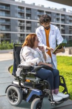Doctor showing medical information on clipboard to woman using electric wheelchair outdoors in a