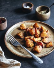 A rustic ceramic plate filled with crispy, herb roasted potatoes sits on a dark table. A fork and