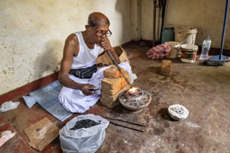 A man sits in a workshop, uses a bellows to start a fire, a man burns sapphires in a small oven in