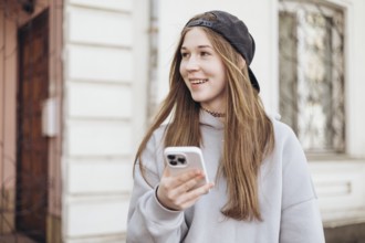Teenage girl in casual attire with a hoodie and cap, smiling while holding a smartphone outdoors.