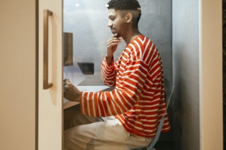 A man wearing a striped sweater works intently in a compact coworking phone booth, representing