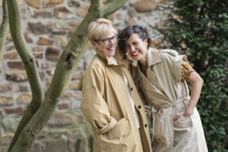 Two fashion women share a joyful moment outdoors, wearing casual trench coats. They laugh and lean