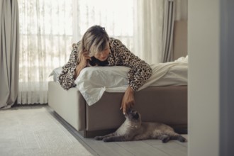 A woman in a stylish leopard-patterned robe leans over her bed to lovingly pet her cat in a cozy