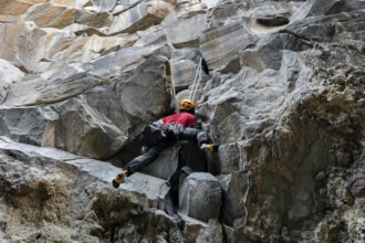 A climber in a red shirt and helmet is scaling a rugged mountain face. The image captures the