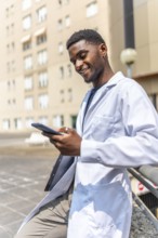 Vertical photo of a cheerful young african doctor using phone during work break outside the