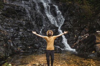 A young man stands with outstretched arms in front of a waterfall in the Choco Andino rainforest in