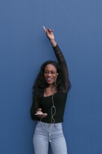 A young woman in casual attire, listening to music with earphones, raises her hand in excitement.