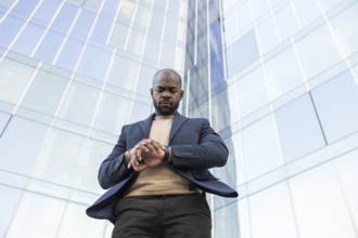 A business cuban man in a suit looks at his watch while standing outside a modern glass skyscraper,