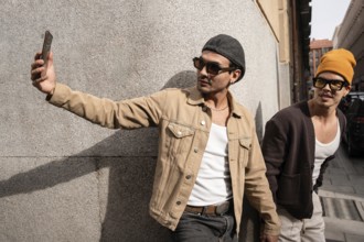 A stylish gay duo takes a selfie against an urban backdrop, dressed in trendy streetwear. One wears