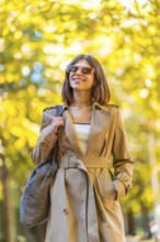 A woman wearing a tan trench coat and sunglasses is walking down a path. She is smiling and holding
