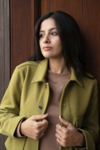 A woman stands against a wooden background, wearing a green jacket and brown top. Her gaze is