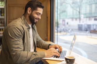 Man smiling and working on a laptop at a wooden table by a window in a cozy coffee shop. A cup of