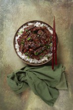 Crispy beef, with green onions and sesame seeds, on a rice pad, in a bowl with chopsticks, on a