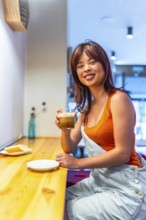 Vertical photo of a young casual chinese smiling woman enjoying morning coffee in a cafeteria