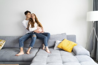 A young couple expecting a baby sits lovingly on a comfortable sofa, embracing each other with