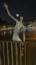 Night view of a sculptural figure on a river with a glowing tethered balloon in the background,