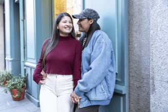 A joyful Latin lesbian couple shares a tender moment outdoors, holding hands and smiling warmly.