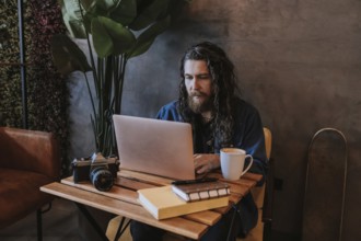 A man with long hair and a beard types on a laptop in a cozy cafe. A vintage camera, books, and a